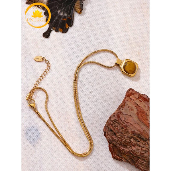 alt="Collier doré avec pendentif en pierre œil de tigre, bijou raffiné posé sur un support naturel"
